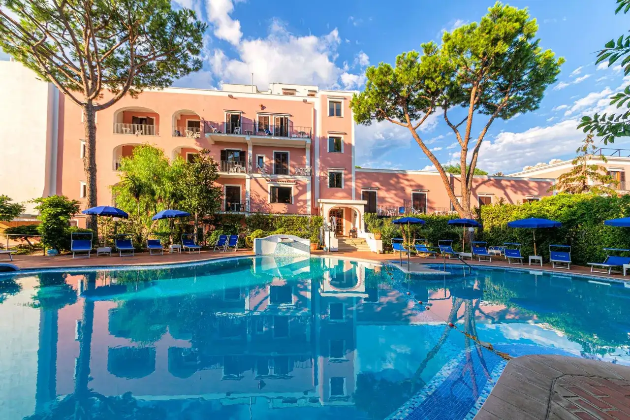 Piscina scintillante con lettini e ombrelloni blu davanti all'hotel Valentino Ischia, immerso nel verde.
