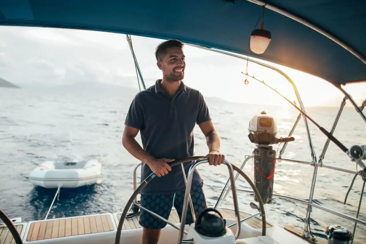 Un homme souriant barre un bateau à voile au coucher du soleil. Un petit bateau gonflable est attaché à l'arrière.