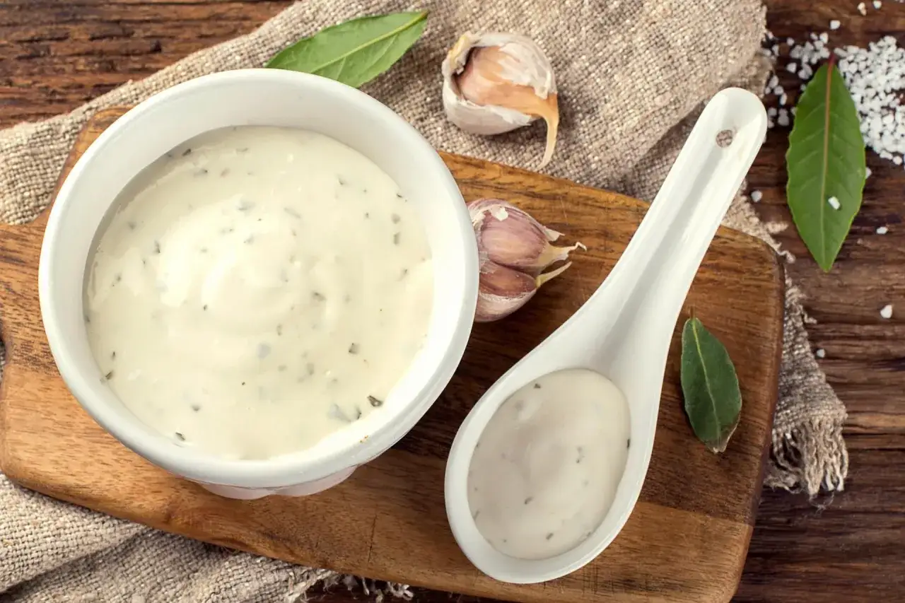 Kremowe aioli w miseczce i na łyżce, obok czosnku i liści laurowych. Idealne do dań.