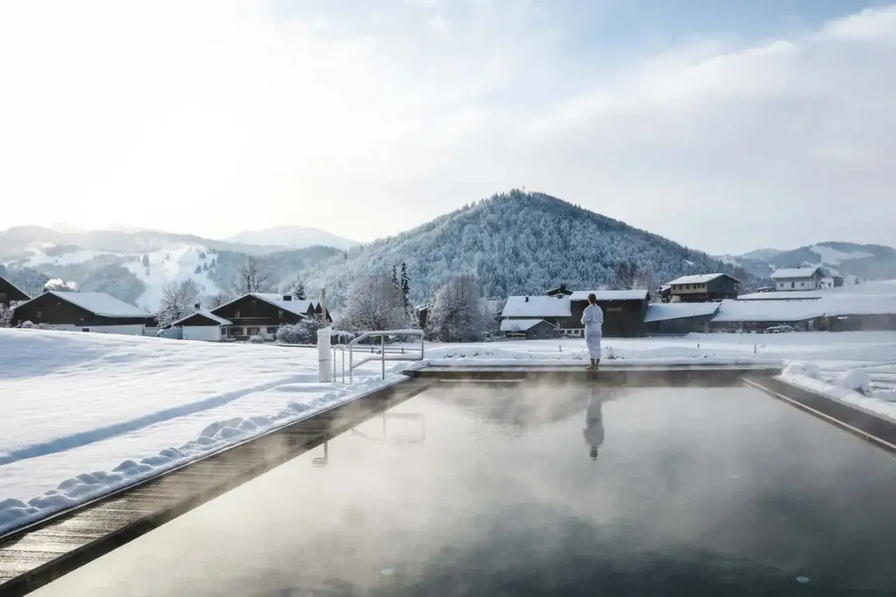 Ein Mann im Bademantel steht am Rand eines dampfenden Pools. Im Hintergrund erstreckt sich eine verschneite Landschaft mit H&auml;usern und einem bewaldeten Berg &ndash; ein idyllisches Bild von Bayern im Winter.