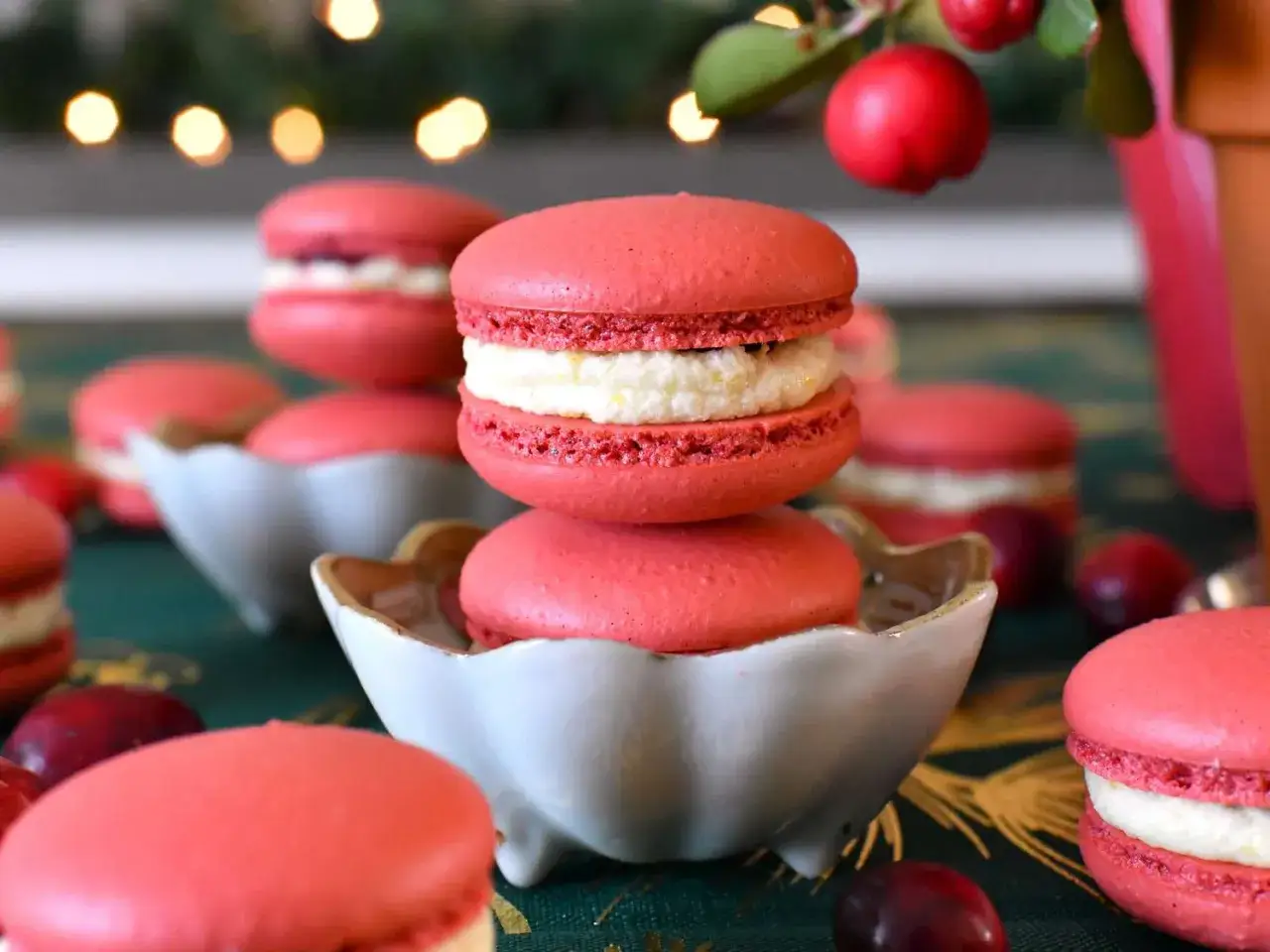 Rosa macarons med vit fyllning, perfekt för ett macarons recept. Dekorerade med röda bär och julbelysning i bakgrunden.