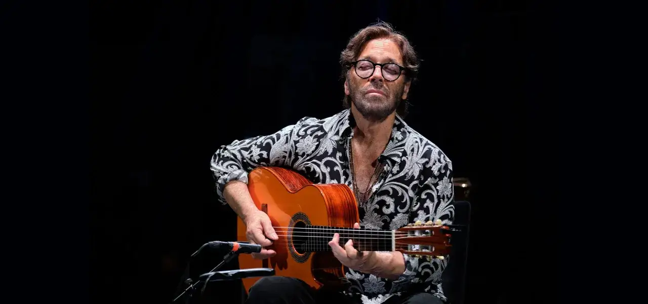 Al Di Meola, z zamkniętymi oczami, gra na gitarze klasycznej w czarno-białej koszuli z wzorem.
