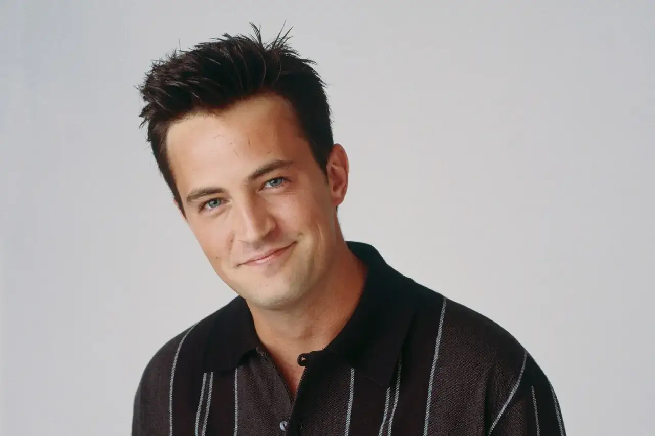 Chandler Bing z uśmiechem na twarzy, w brązowej koszulce w paski.