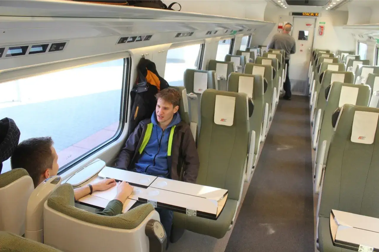 Wnętrze wagonów Pendolino. Dwóch młodych mężczyzn siedzi przy stoliku, rozmawiając. W tle widać rzędy zielonych foteli.