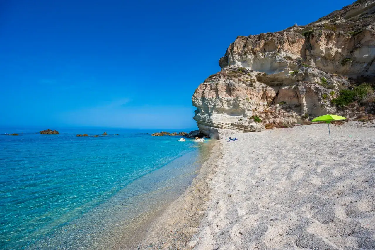 A secluded beach in Calabria, Italy, with crystal-clear turquoise water, white sand, and dramatic cliffs. A bright green umbrella offers shade on this idyllic scene.