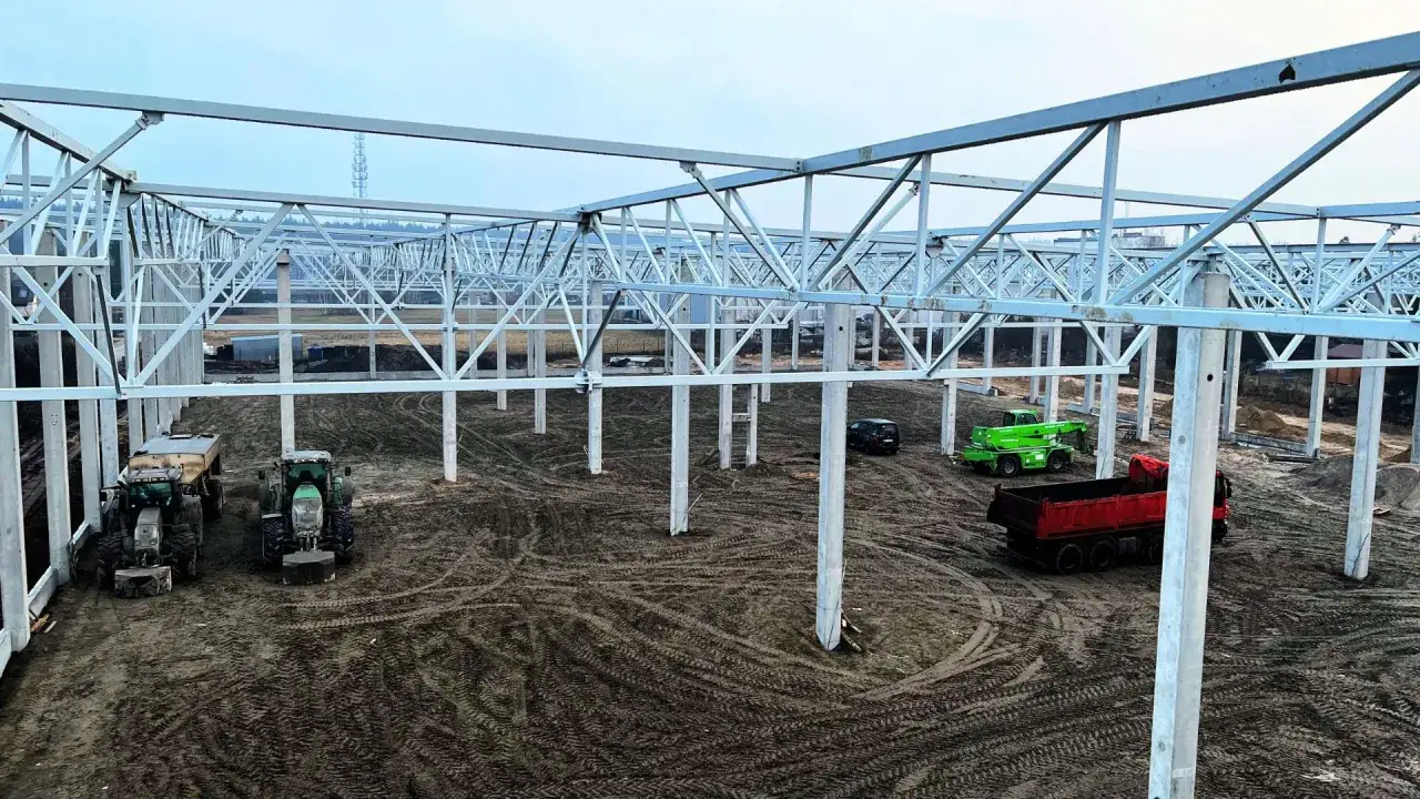 Trwa budowa hali produkcyjnej. Na placu budowy stoją traktory, zielony podnośnik, czerwona wywrotka i czarny samochód.