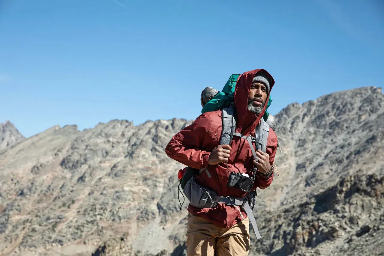 Mann mit Rucksack und Kamera in den Bergen. Er tr&auml;gt eine rote Jacke und eine graue M&uuml;tze &ndash; so zieht man zum Wandern an.