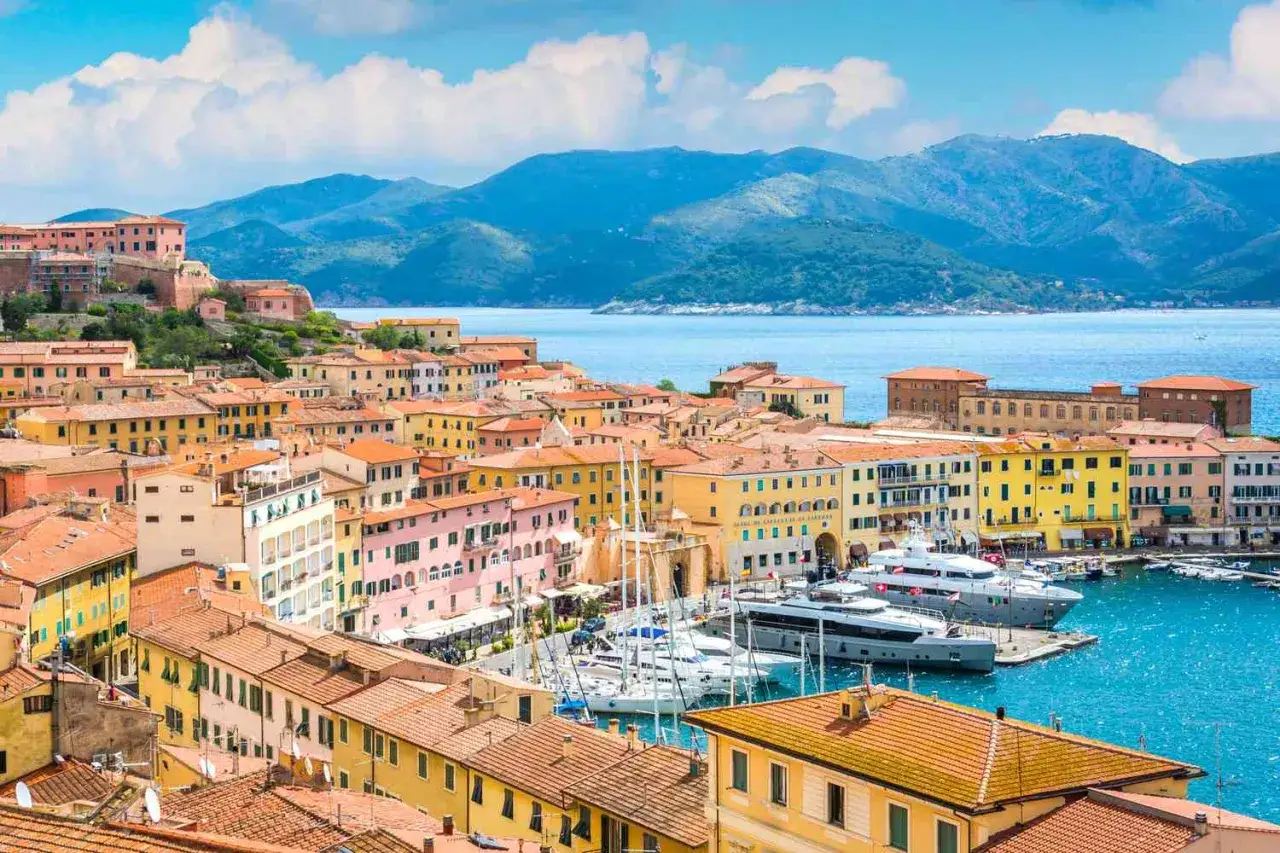 F&auml;rgstarka hus vid en hamn p&aring; Elba, Italien. Bl&aring;tt hav och gr&ouml;na berg i bakgrunden.