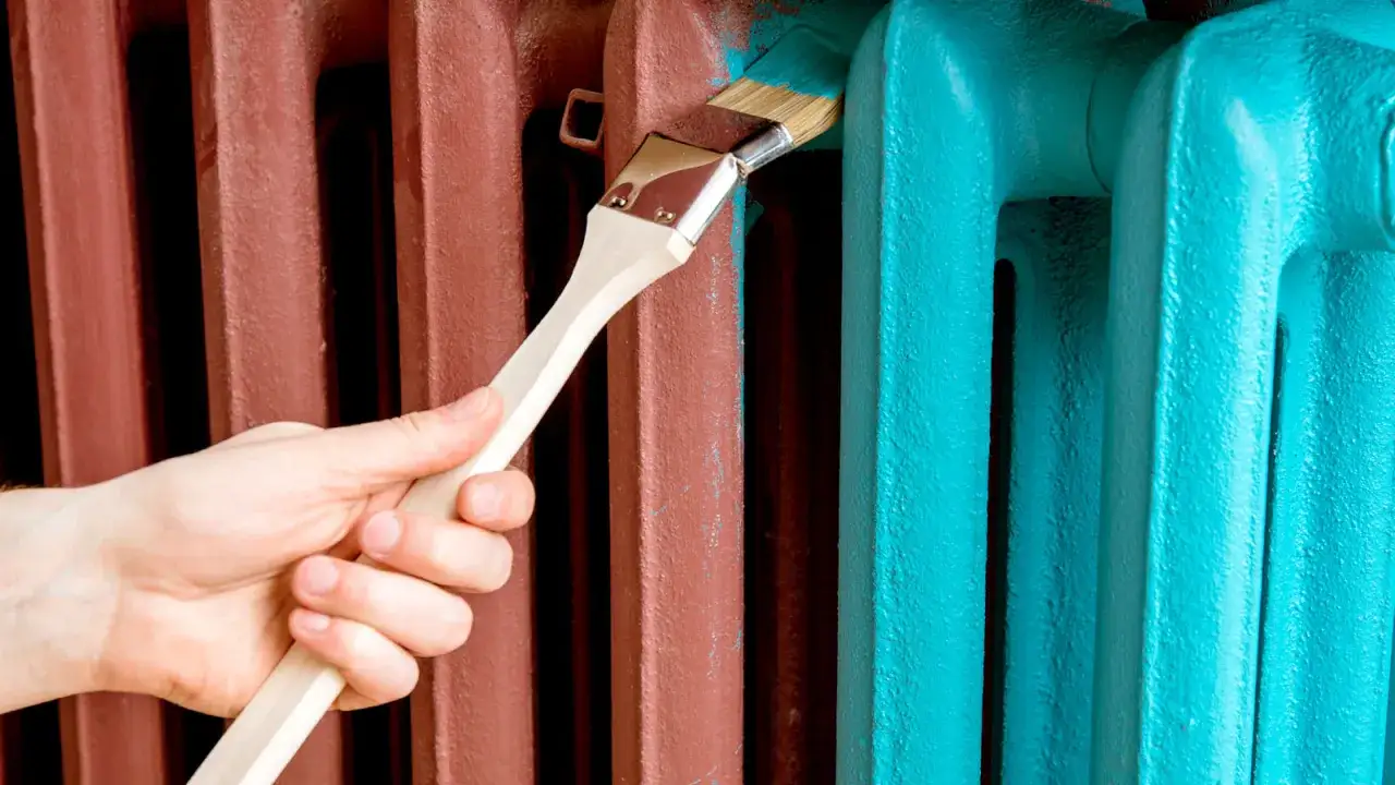 Mano pintando un radiador de color marrón rojizo con pintura azul turquesa. El proceso de pintar radiadores puede transformar un espacio.