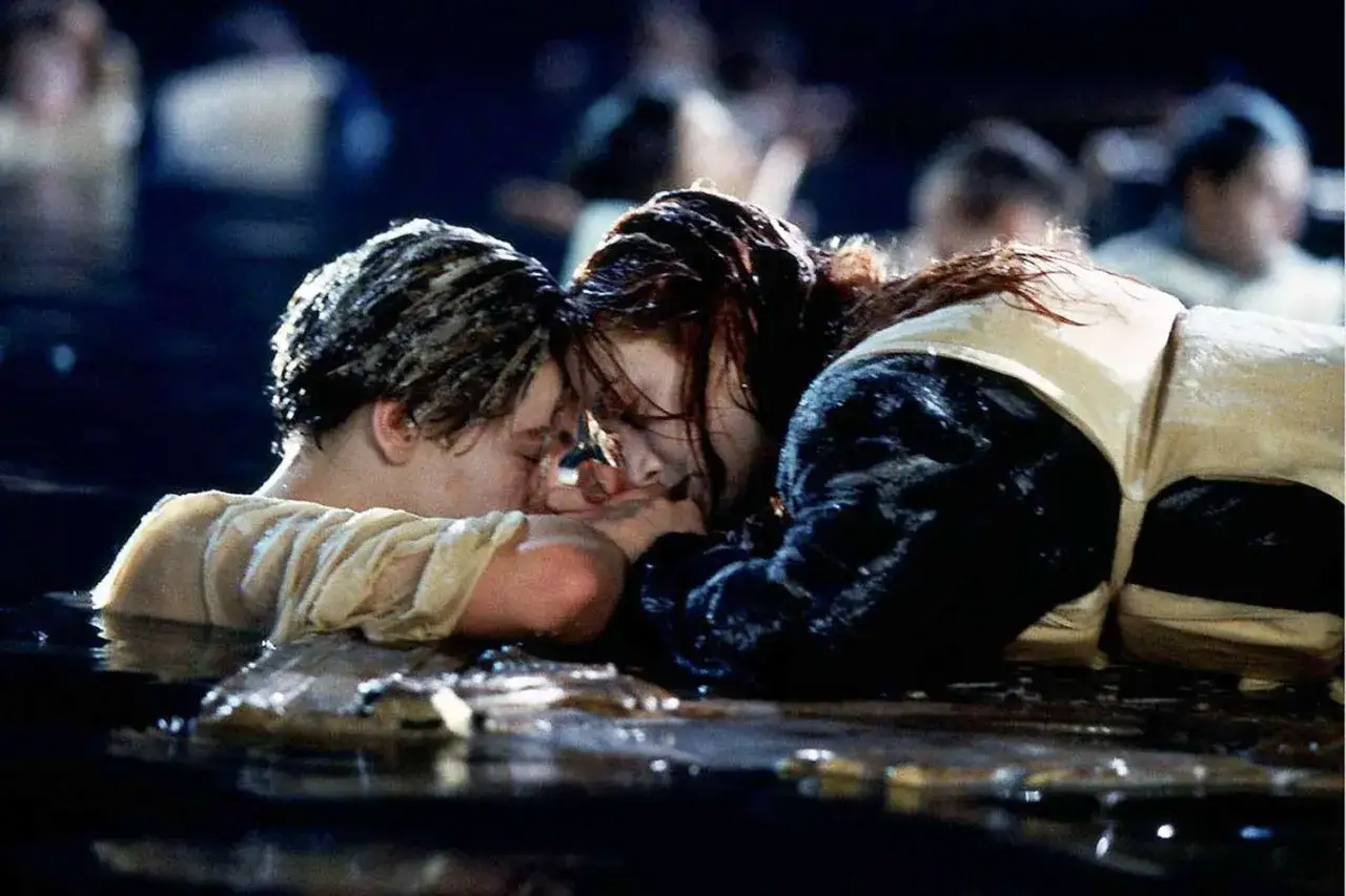 Jack i Rose, główni bohaterowie filmu Titanic, w dramatycznej scenie. Ich twarze są blisko siebie, w wodzie, w tle widać innych pasażerów.