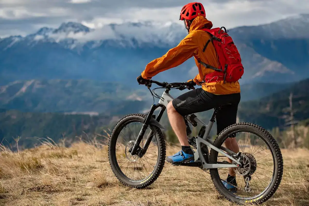 A mountain biker in an orange jacket and red helmet, with a red backpack, rides a full-suspension bike on a grassy ridge. These are essential mtb must haves for an epic adventure.
