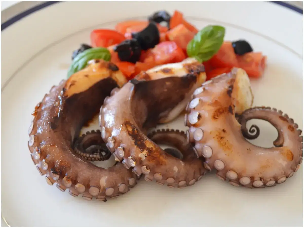 Tentacoli di polpo alla griglia, succulenti e dorati, serviti con pomodorini freschi, olive nere e foglie di basilico. Un piatto che profuma di mare.
