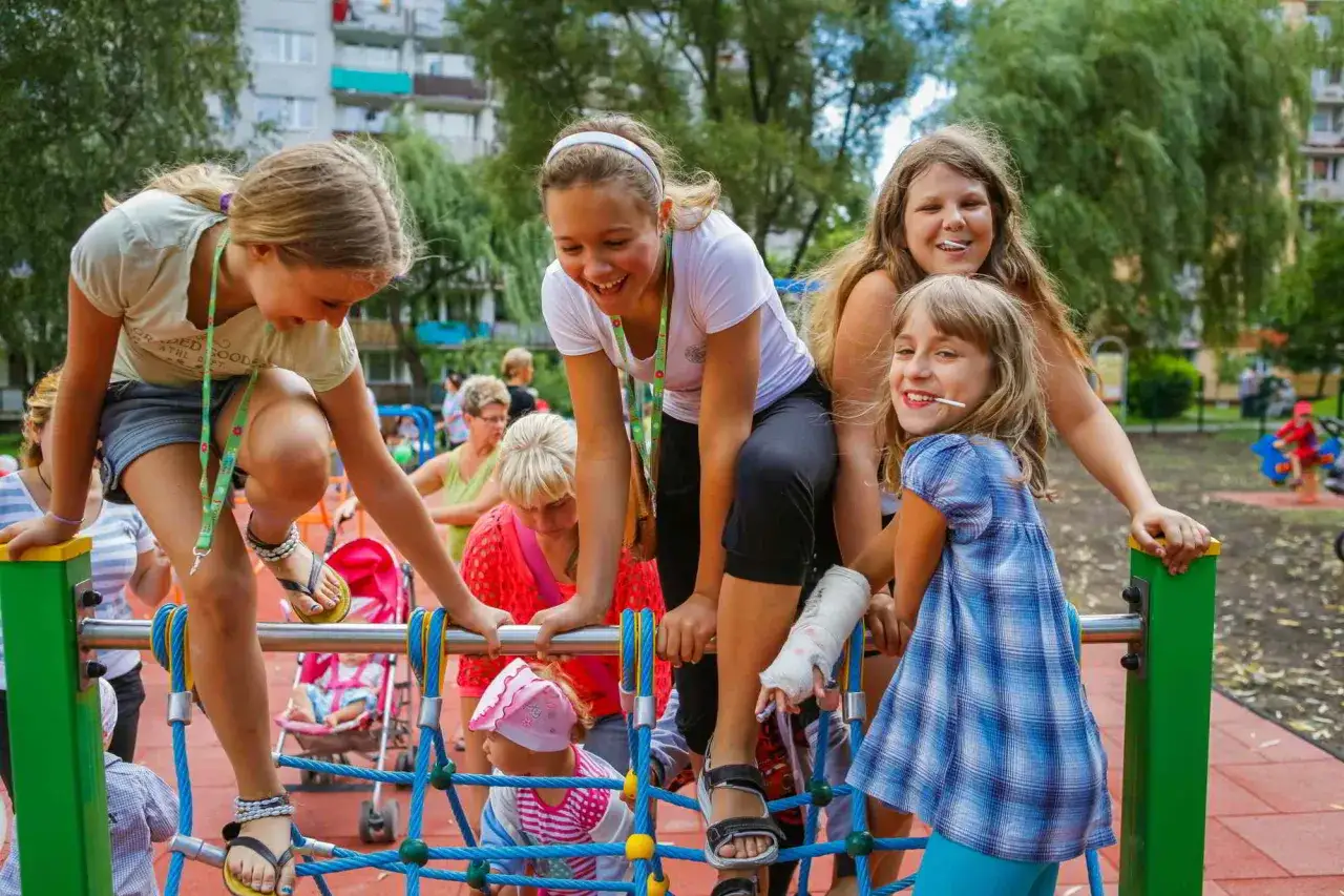 Radosne dzieciaki na placu zabaw, jak wujek mówi na dziecko, śmieją się i wspinają. Jedna z dziewczynek ma rękę w gipsie.