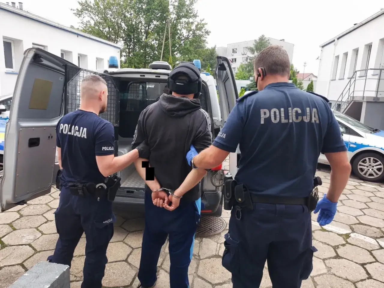 Policja doprowadzenie do zakładu karnego. Dw&oacute;ch funkcjonariuszy w mundurach eskortuje zatrzymanego w kajdankach do policyjnego vana.