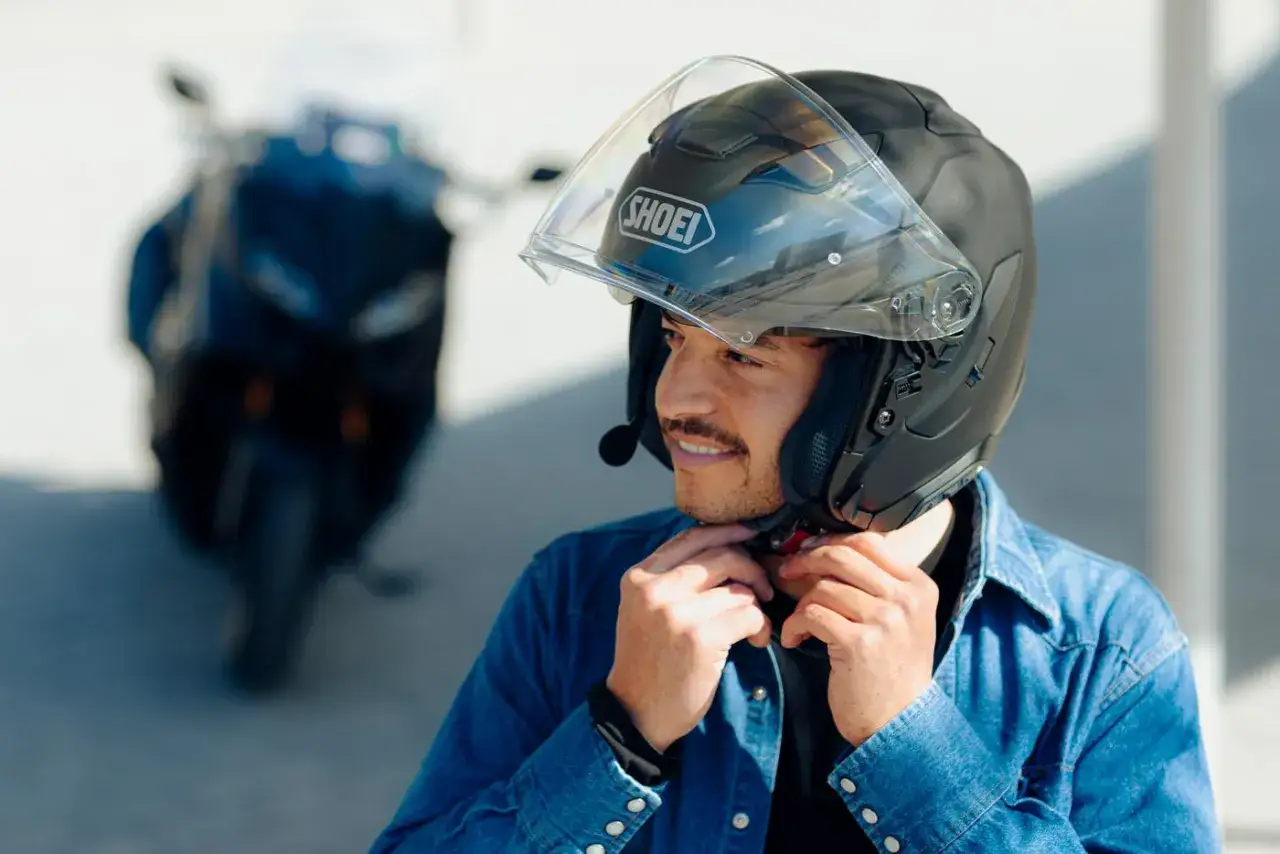 Hombre ajustándose un casco jet negro Shoei, listo para cumplir la normativa. Detrás, una moto oscura.