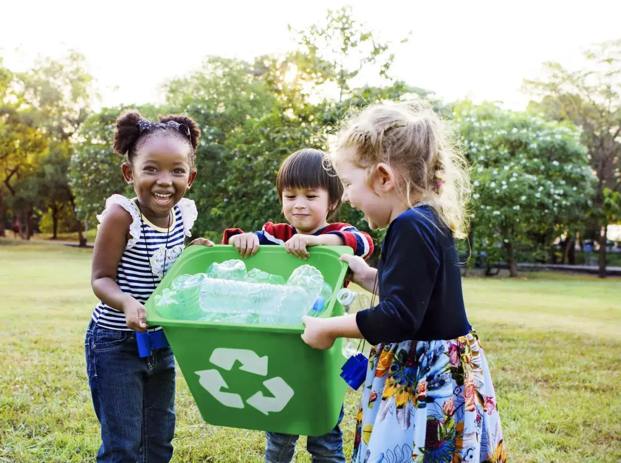 Niños sonriendo mientras reciclan botellas de plástico en un parque. Actividades medio ambiente infantil para un futuro mejor.