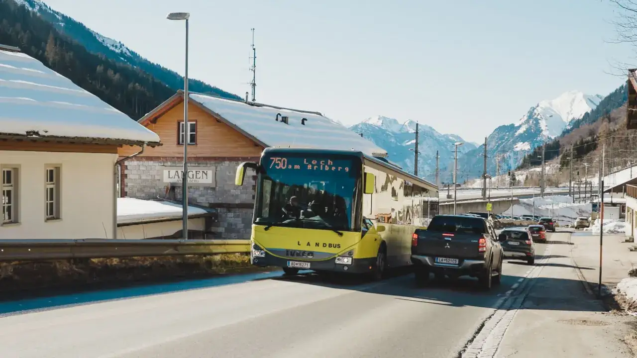 Gelber Landbus f&auml;hrt Richtung Lech am Arlberg. Schneebedeckte Berge im Hintergrund, Langen am Berg im Vordergrund.