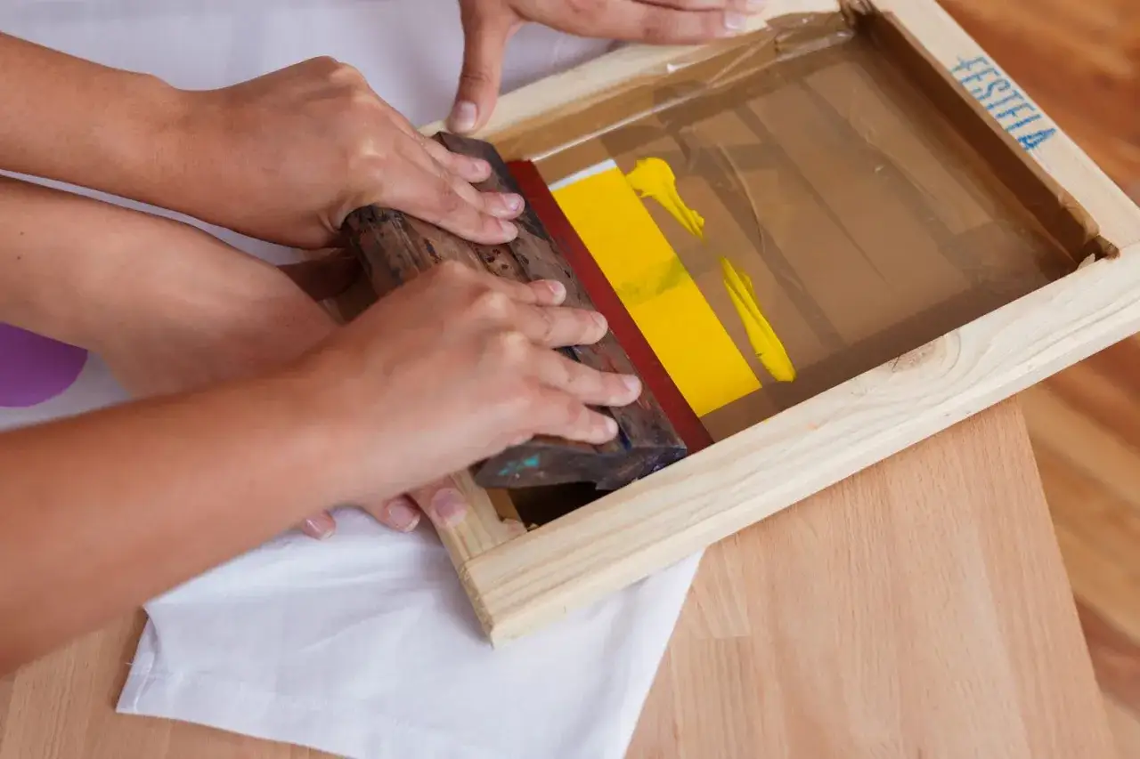 Manos aplicando tinta amarilla con rasqueta sobre pantalla para serigrafía en casa.