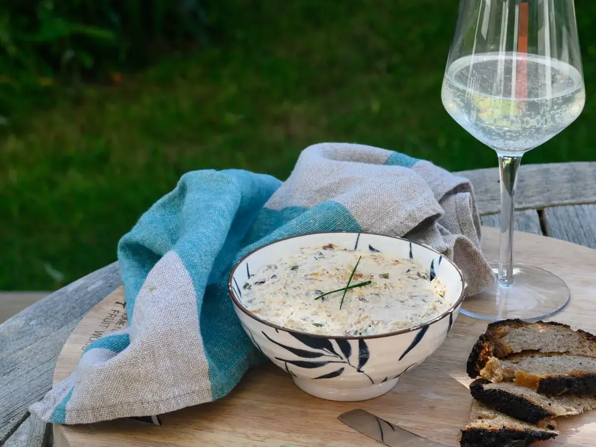 Un bol de rillettes de maquereau garni de ciboulette, servi avec du pain grillé et un verre de vin blanc pétillant, sur une table en bois en extérieur.