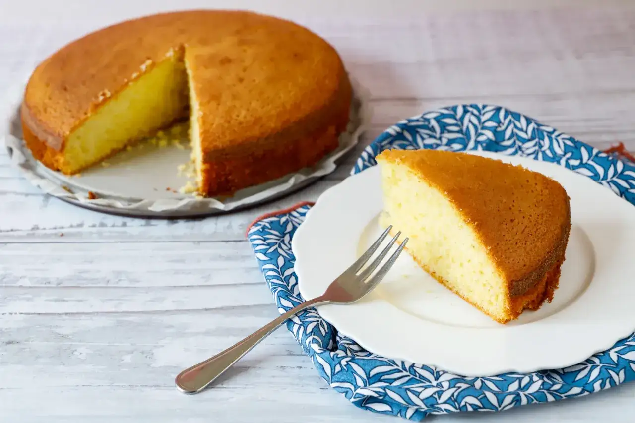 Una fetta di torta senza lattosio dorata e soffice su un piatto bianco, accanto a una torta intera su un vassoio.