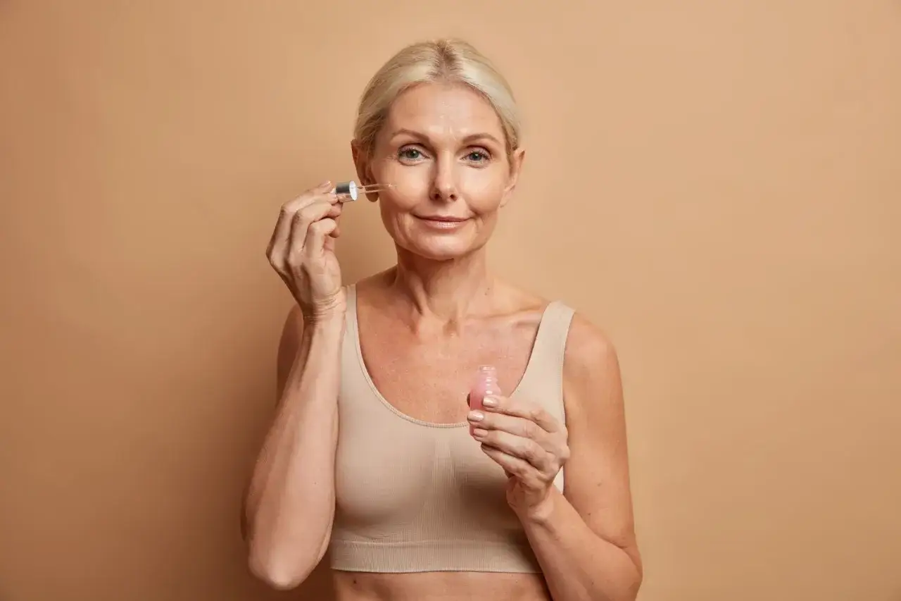 Kobieta z blond włosami aplikuje serum na twarz, dbając o sk&oacute;rę w okresie menopauzy.