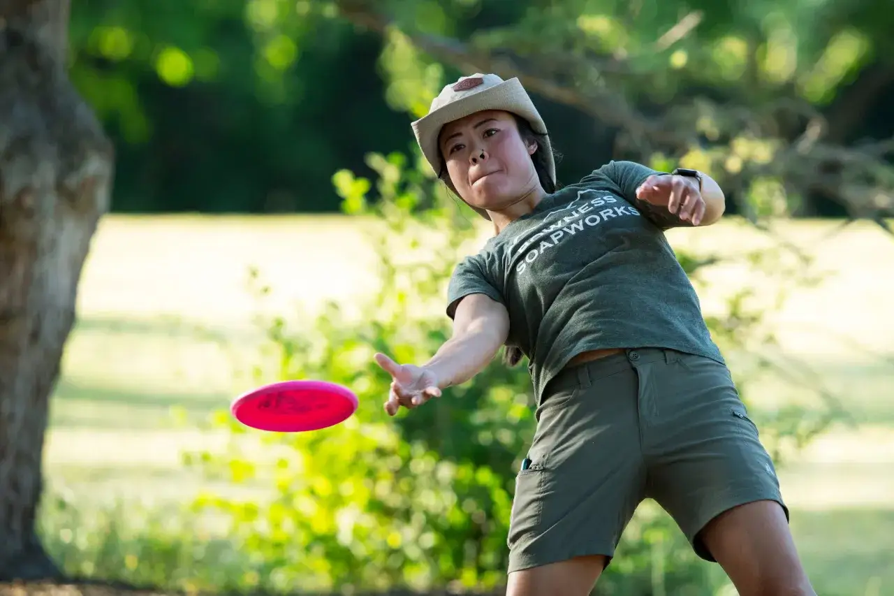 Une joueuse de disc golf lance un frisbee rose vif dans un parc ensoleill&eacute;.