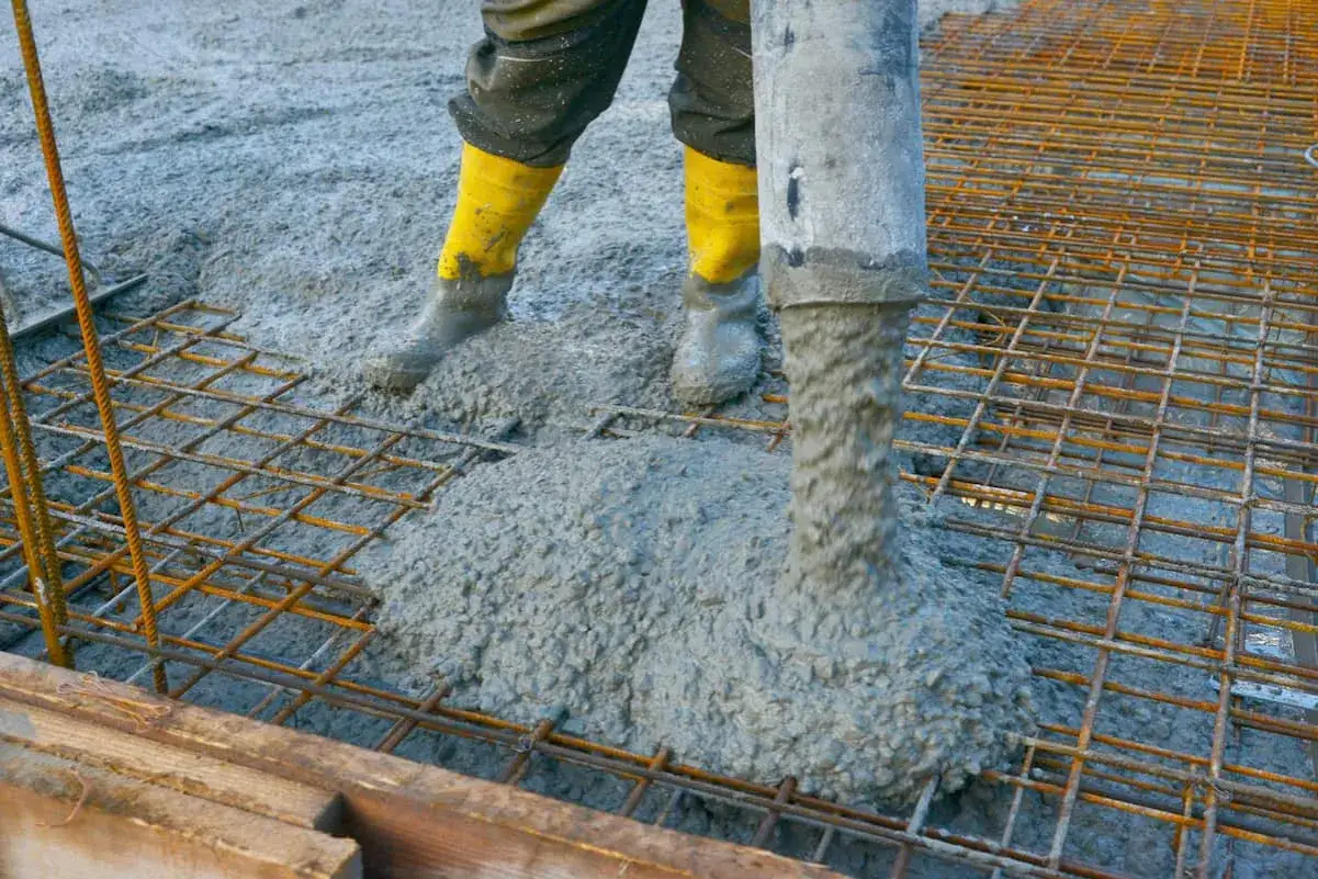 Beton flie&szlig;t &uuml;ber Stahlmatten. Arbeiter in gelben Stiefeln &uuml;berwachen den Prozess, w&auml;hrend der fl&uuml;ssige Beton die Gitterstruktur bedeckt.