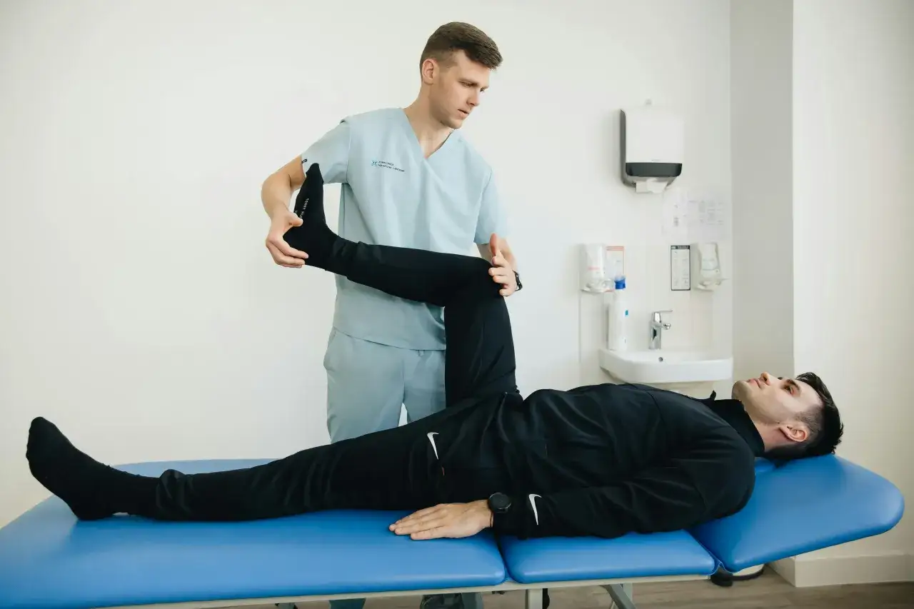 Fysioterapeut hj&auml;lper patient att stretcha ischias. Patienten ligger p&aring; en bl&aring; brits medan fysioterapeuten lyfter patientens ben.