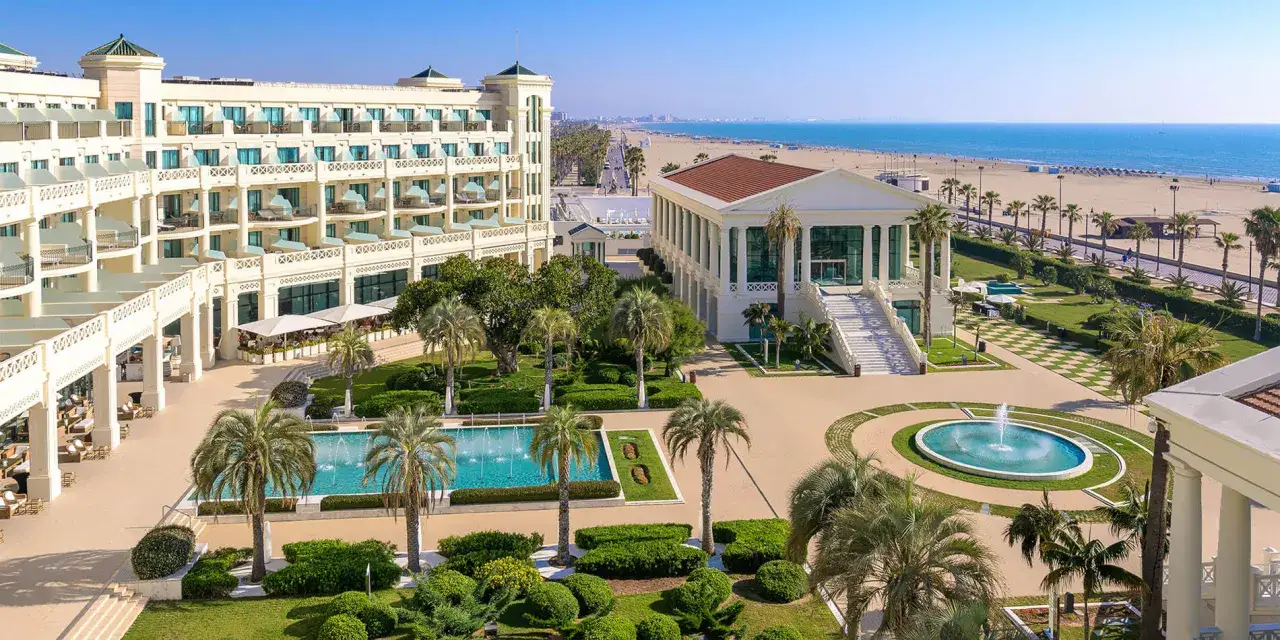 El balneario de la Alameda, un hotel de lujo frente al mar, con piscinas, jardines y palmeras, se extiende hacia una playa dorada y el mar azul.