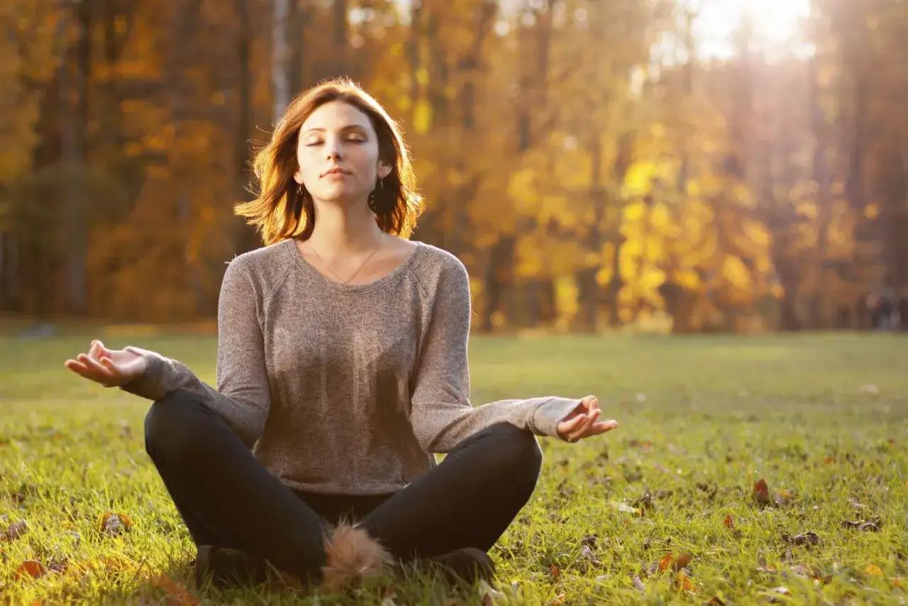A woman meditates in a park, finding her inner calm and learning how to be at peace amidst the golden autumn foliage.