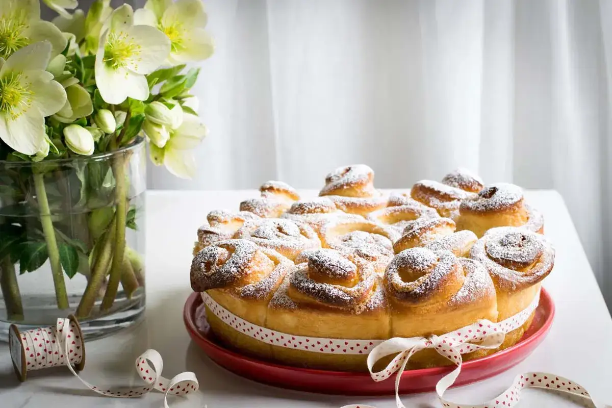 Una deliziosa torta delle rose, cosparsa di zucchero a velo, decorata con un nastro a cuoricini, accanto a un vaso di fiori bianchi.