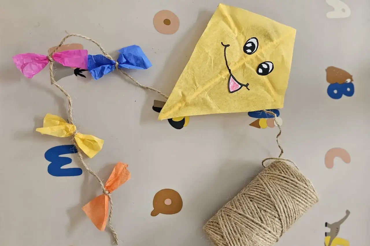 Ein gelber Papierdrachen mit lustigem Gesicht und bunten Papierbanderolen h&auml;ngt an einer Schnur. Perfekt zum **Drachen basteln**!