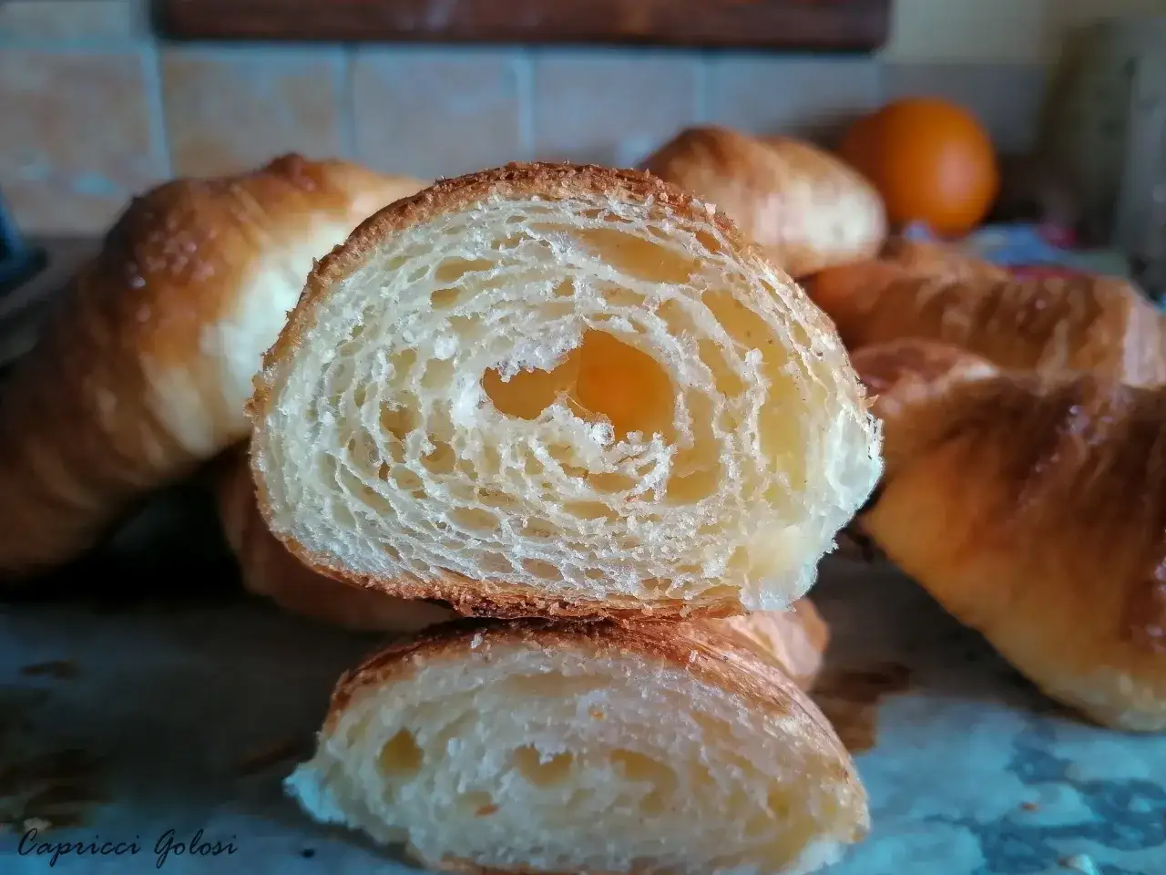 Cornetti con lievito madre, sfogliati e dorati, con un interno alveolato e soffice. Un'arancia sullo sfondo suggerisce un tocco di freschezza.