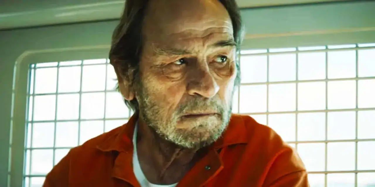 Tommy Lee Jones w pomarańczowym kombinezonie, zamyślony, z siwą brodą, na tle kratowanego okna.