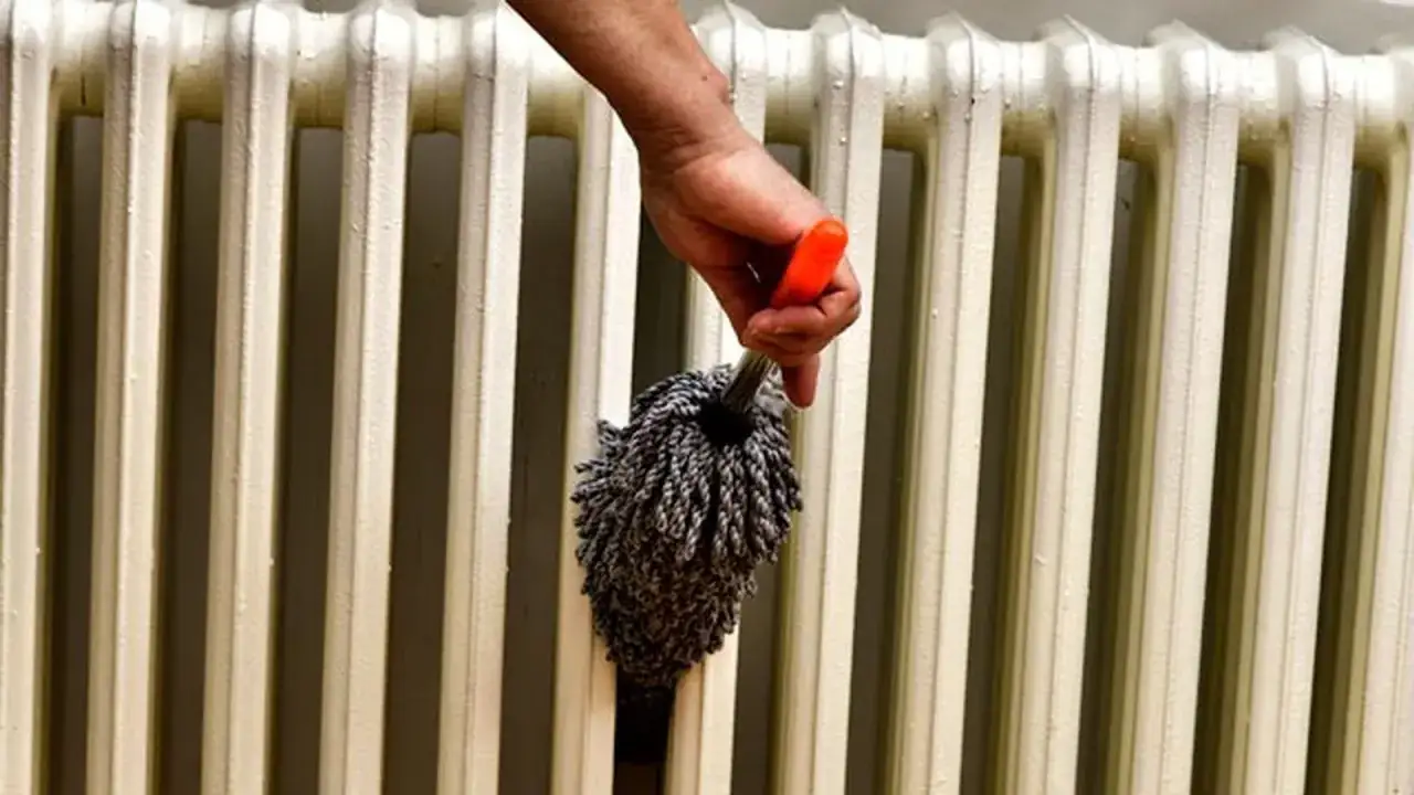Mano con plumero limpiando un radiador blanco. Una gu&iacute;a visual para como limpiar radiador y mantener tu hogar c&aacute;lido y limpio.