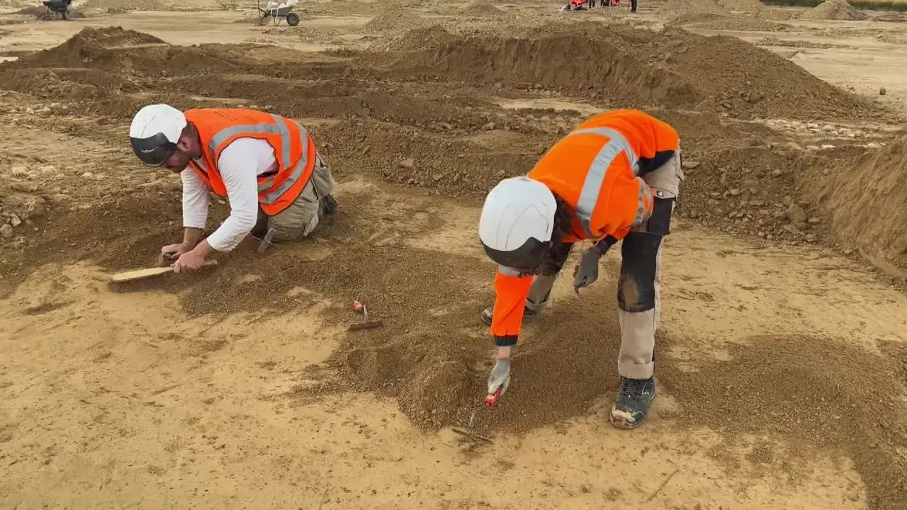 Deux arch&eacute;ologues, &eacute;quip&eacute;s de casques et de gilets de haute visibilit&eacute;, travaillent avec minutie sur un site de fouilles. Une **d&eacute;couverte arch&eacute;ologique** se profile sous leurs outils.