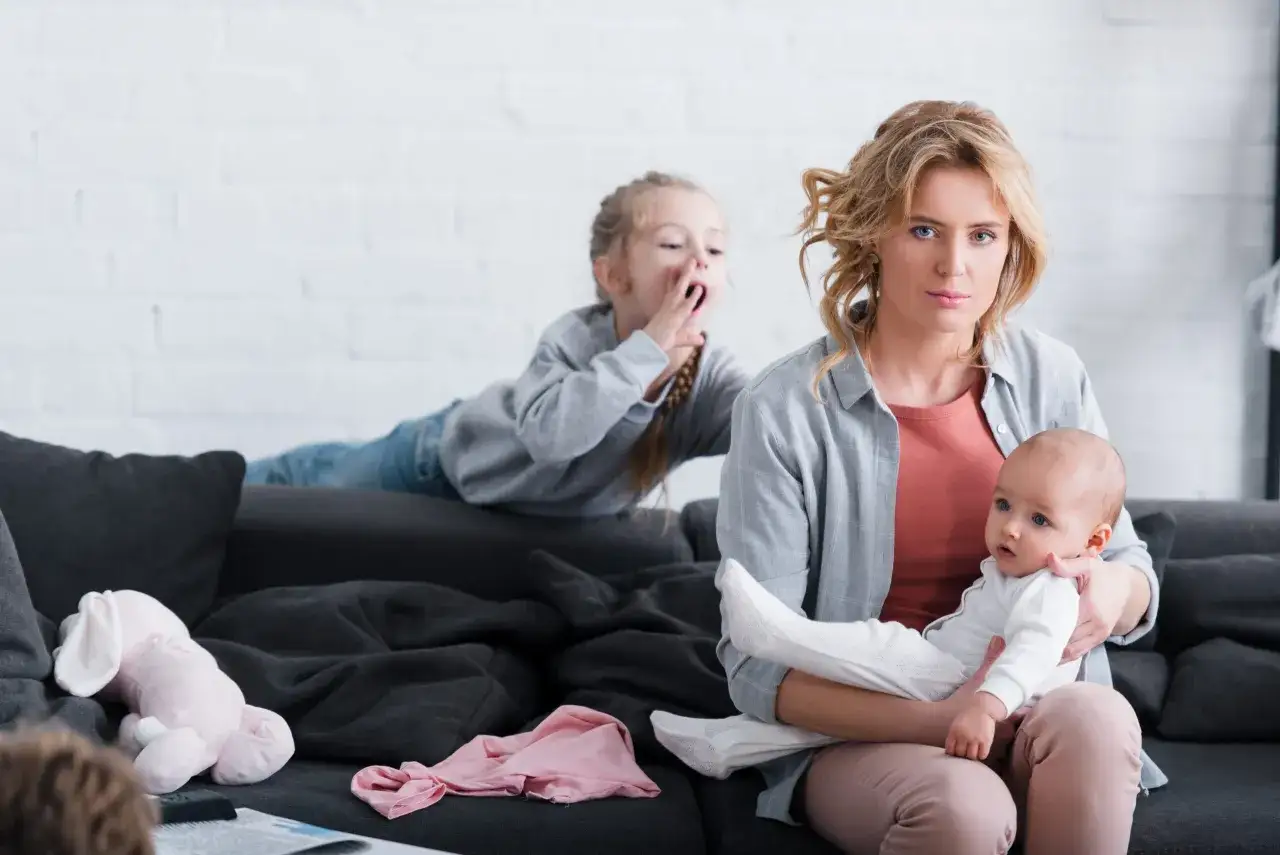 Mutter mit Baby auf dem Arm, ältere Tochter gähnt. Die narzisstische Mutter wirkt abwesend, während die Kinder ihre Aufmerksamkeit fordern.