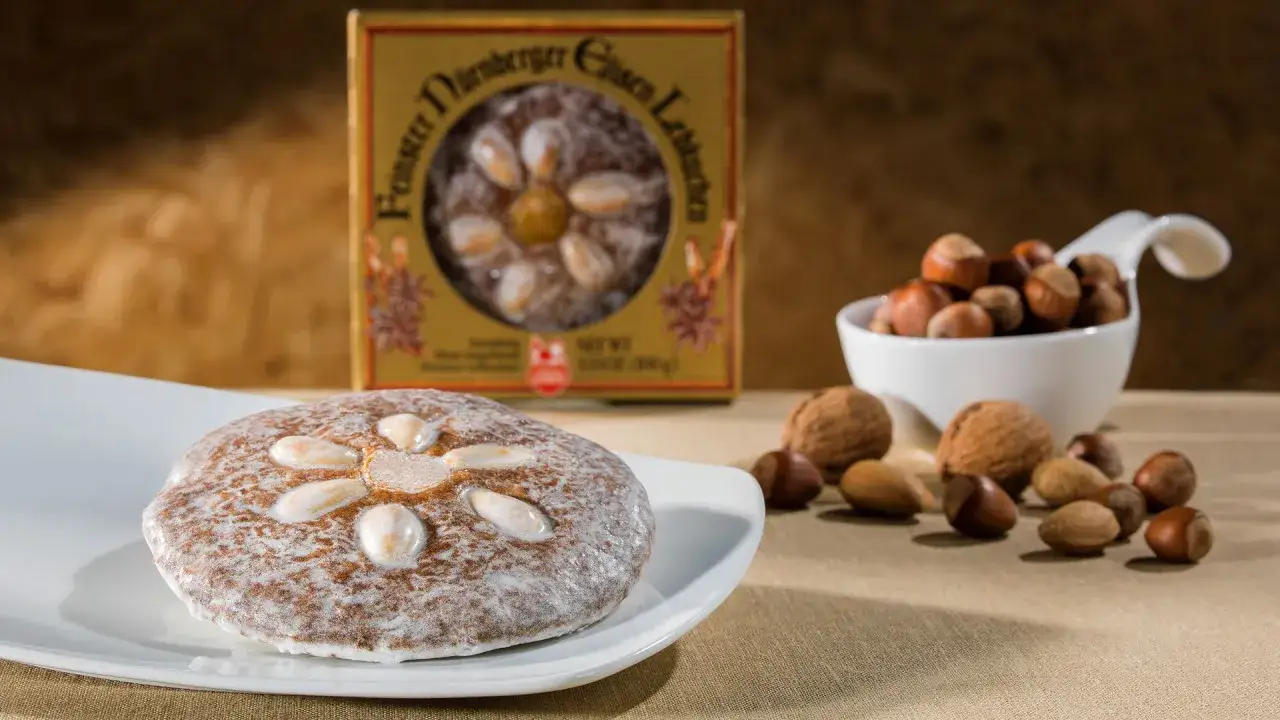 Ein Nürnberger Elisenlebkuchen, verziert mit Mandeln, liegt auf einem Teller. Daneben eine Schale mit Haselnüssen und Walnüssen. Der beste Elisenlebkuchen für gemütliche Stunden.