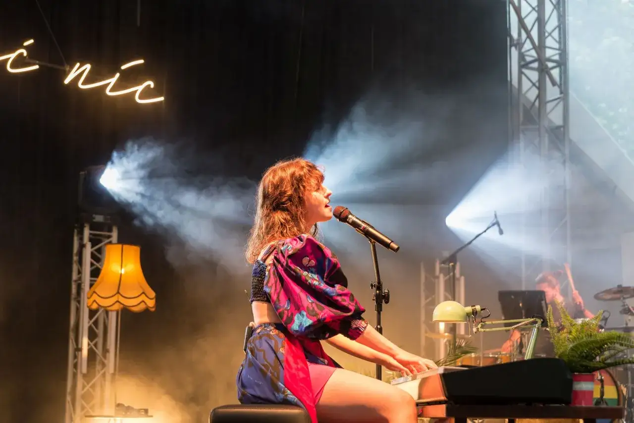 Kasia Sienkiewicz gra na keyboardzie i śpiewa do mikrofonu, otoczona dymem scenicznym i światłami.