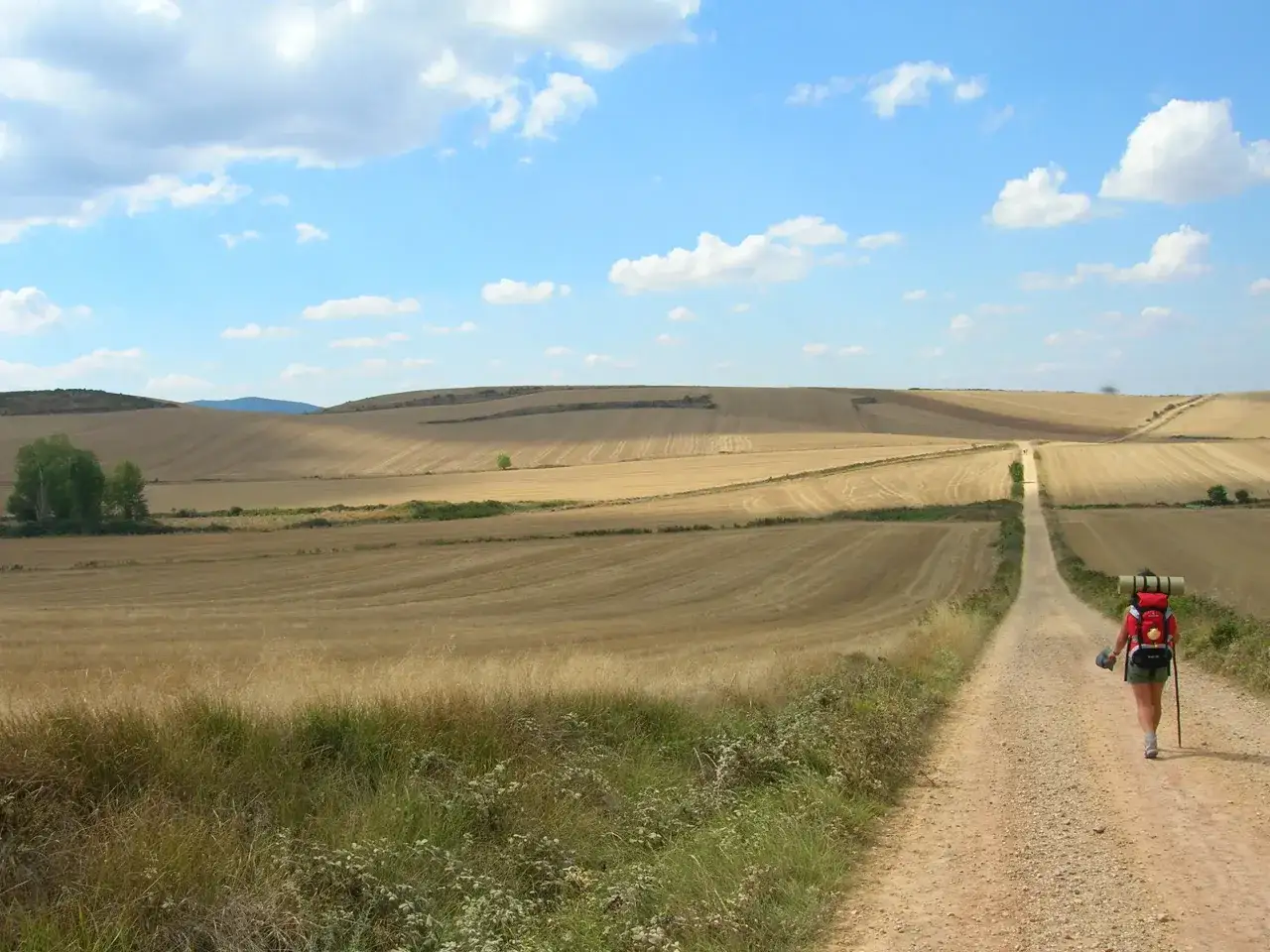 Peregrina con mochila roja camina por un sendero rural, parte del camino de Santiago desde Madrid, bajo un cielo azul con nubes blancas.