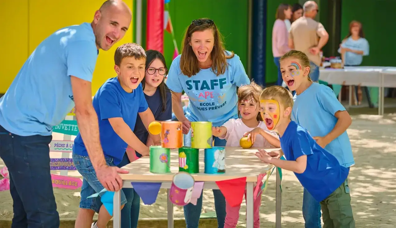 Des enfants et des adultes s'amusent à un jeu de lancer de balles sur des boîtes décorées. C'est ça une kermesse : joie, jeux et convivialité !
