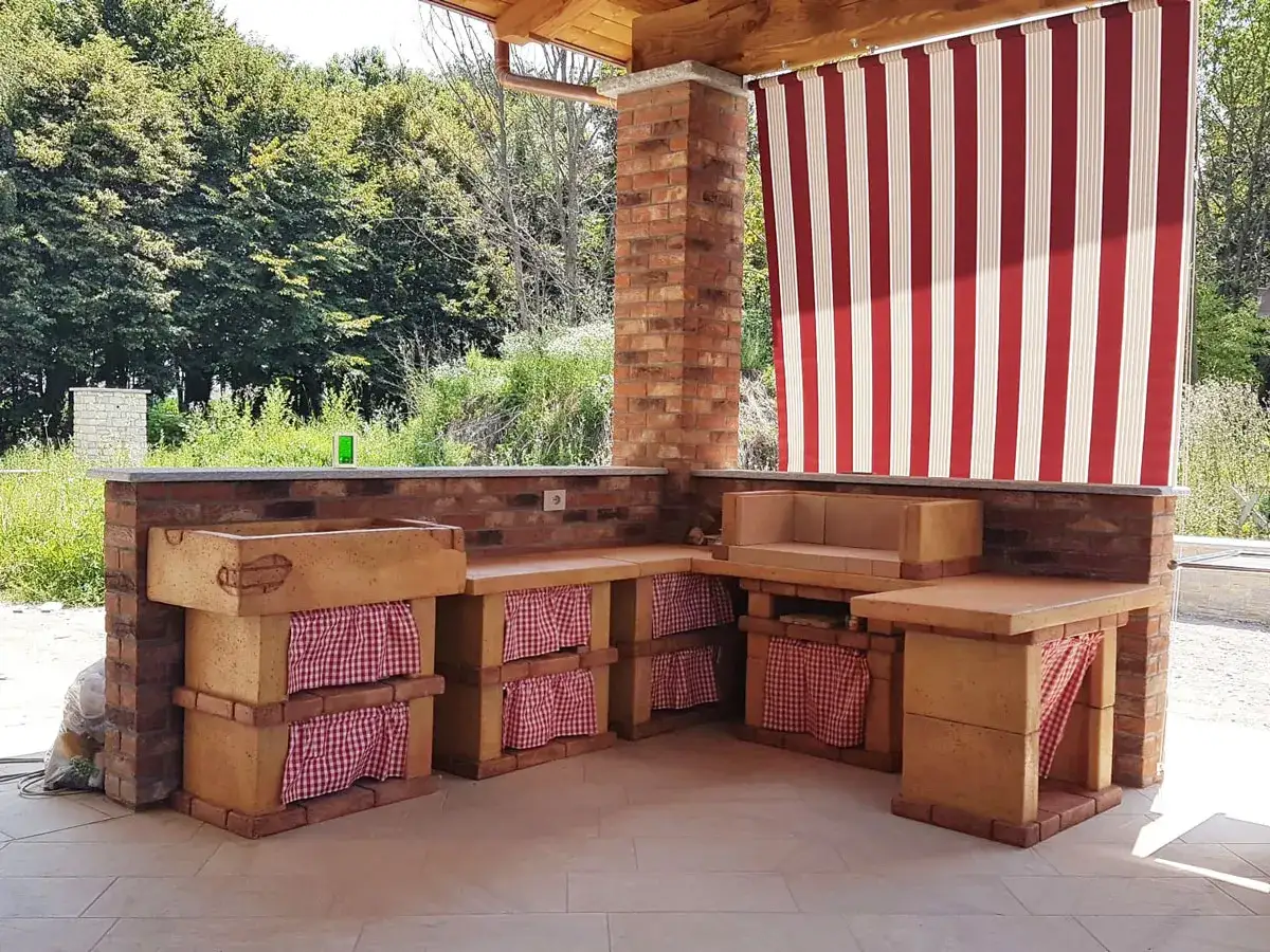 Una splendida cucina all'aperto fai da te con lavello in pietra, piano di lavoro e seduta in mattoni, decorata con tende a righe rosse e bianche.