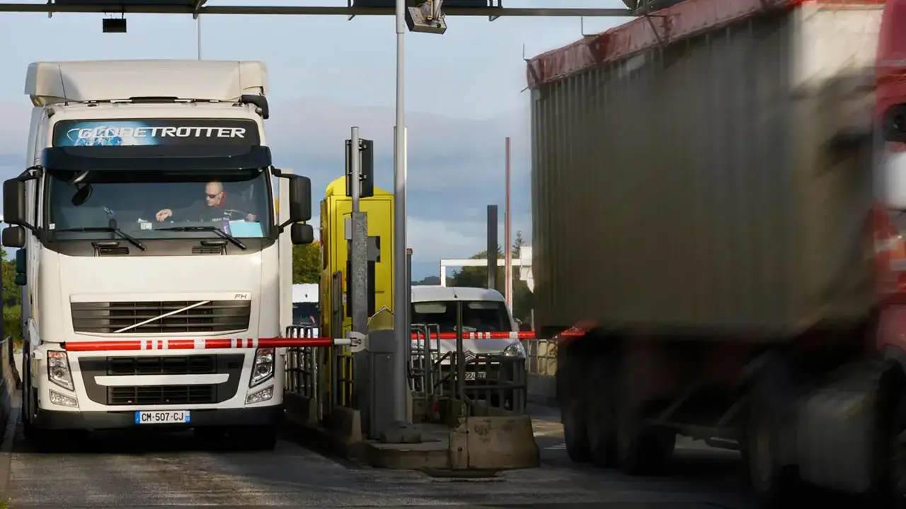 Cami&oacute;n blanco en peaje, listo para cruzar a Luxemburgo o Holanda. Otro cami&oacute;n pasa borroso.