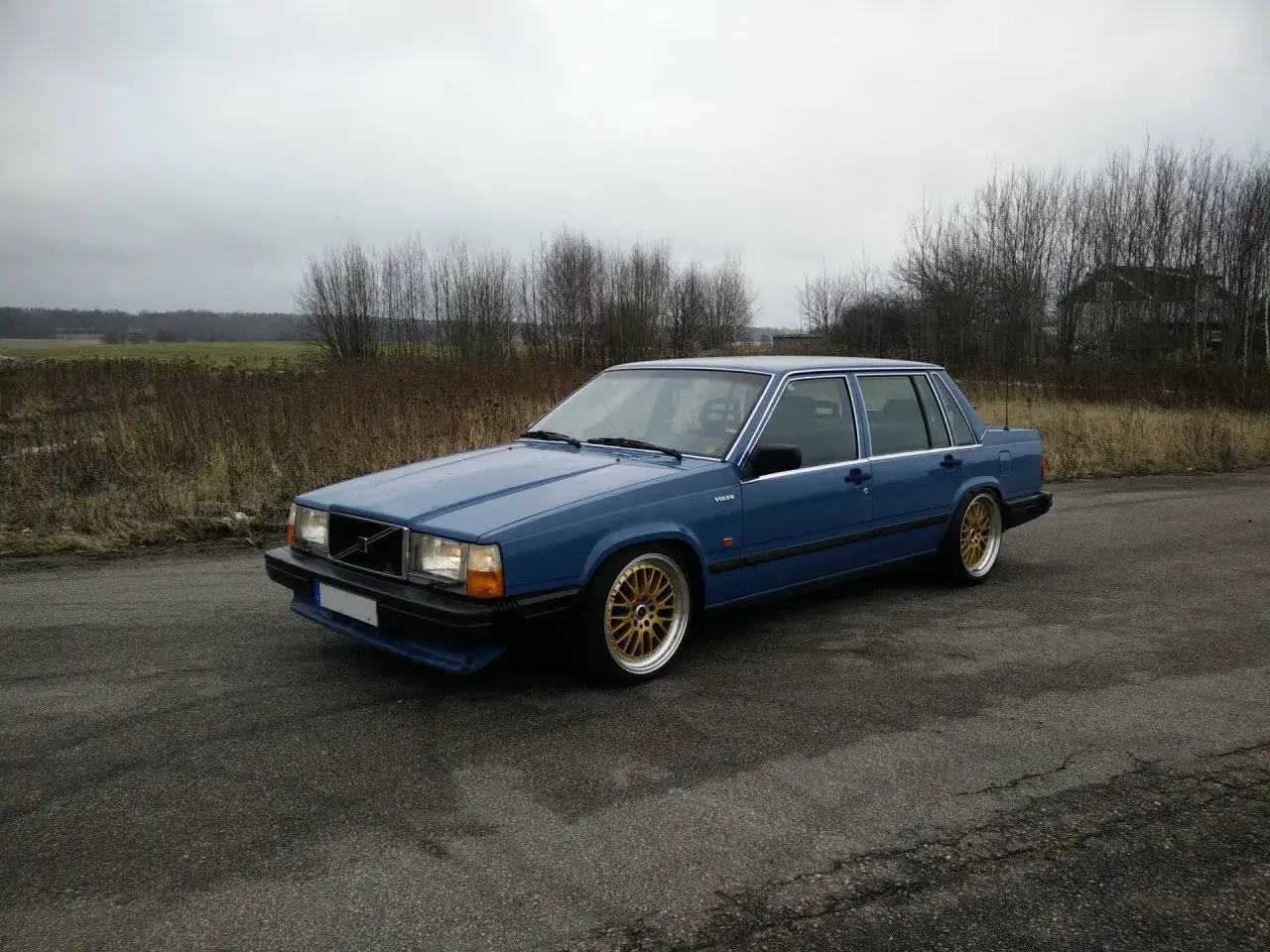 Bl&aring; Volvo 740 fr&aring;n 1990 med guldf&auml;rgade f&auml;lgar st&aring;r parkerad vid en grusv&auml;g i ett &ouml;de landskap.