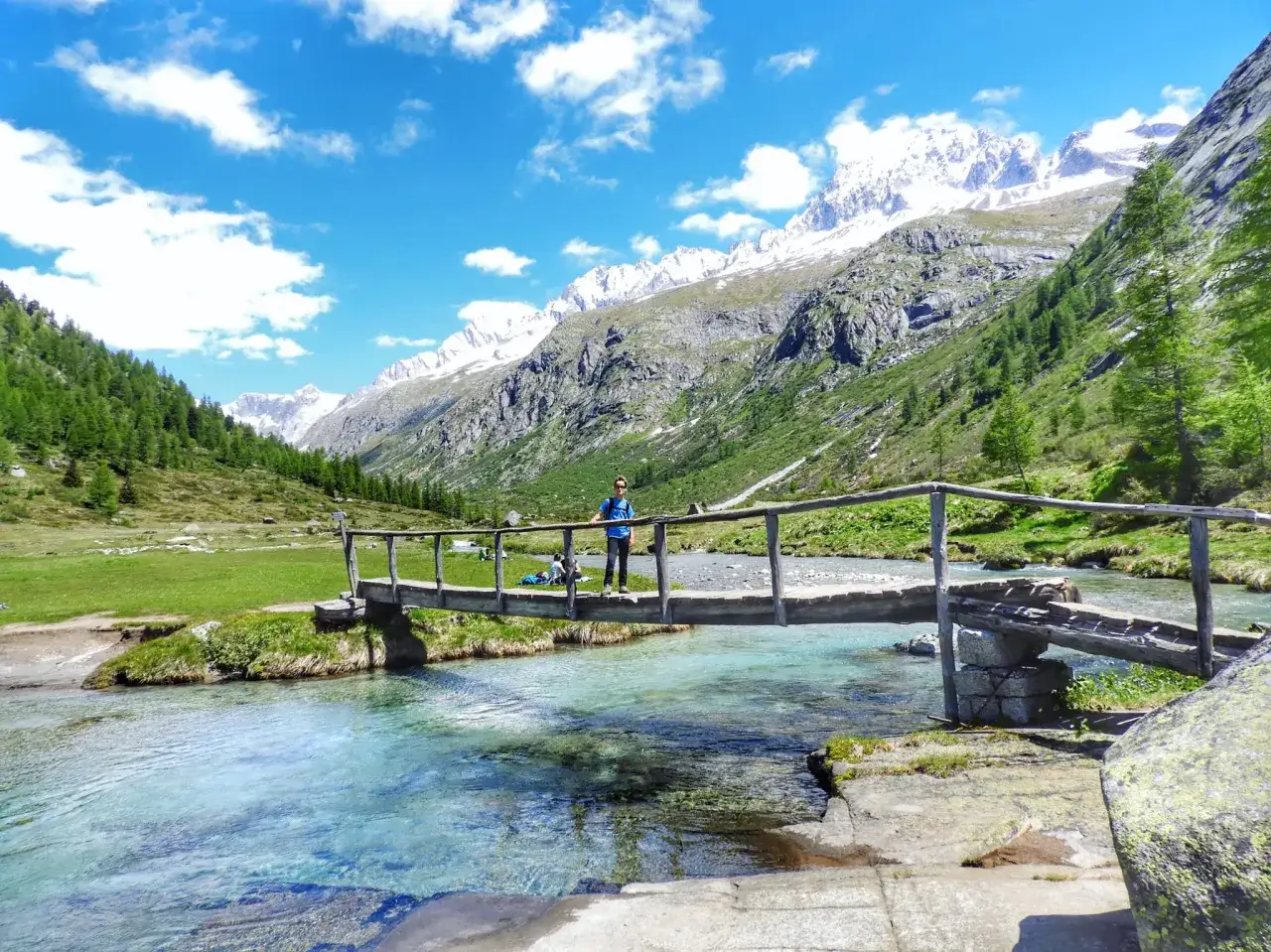 Un uomo su un ponte di legno nel **Val di Fumo**, con montagne innevate sullo sfondo e un fiume cristallino.
