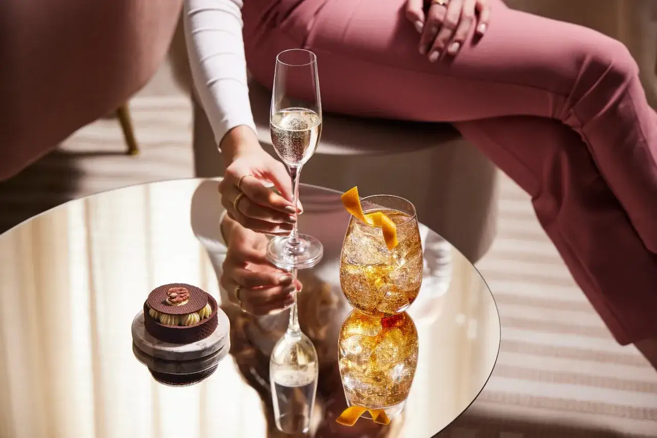 Elegancki prosecco drink i deser na lustrzanym stoliku. Kobieta w różowych spodniach sięga po kieliszek musującego napoju.