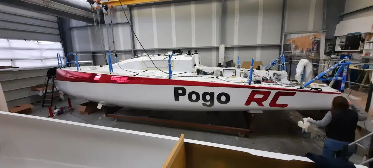 Un voilier Pogo RC blanc et rouge est en cours de finition dans un atelier. Une personne nettoie la coque.