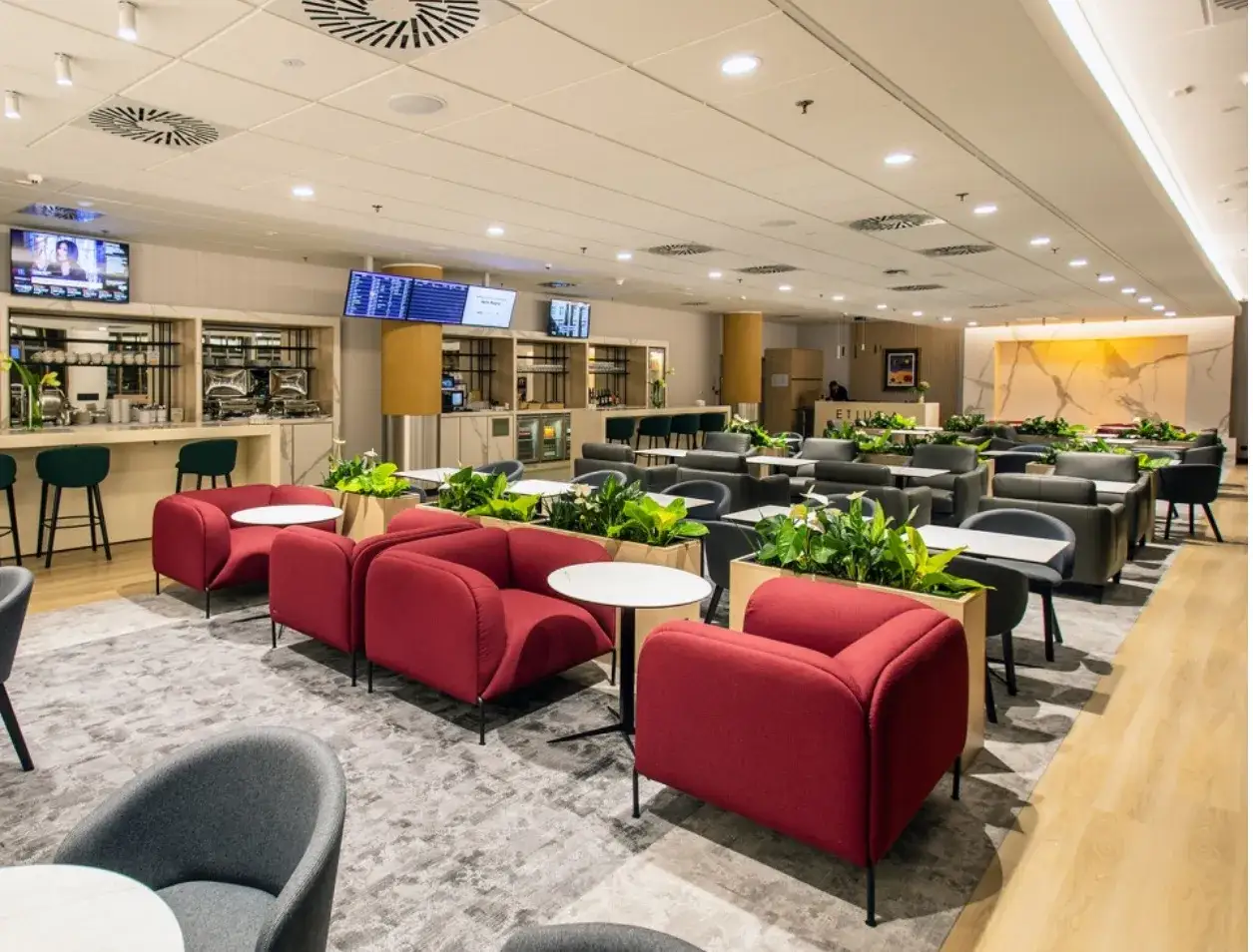 Elegancka poczekalnia z wygodnymi fotelami w kolorze czerwonym i szarym, stołami, roślinami i barem. Idealne miejsce na relaks przed podróżą, jak w strefie Santander Select.