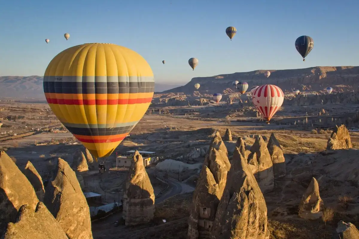Fler än tio luftballonger svävar över Cappadocias unika landskap. En stor gul ballong med färgglada band dominerar förgrunden. Detta är en fantastisk vy av fakta om Turkiet.