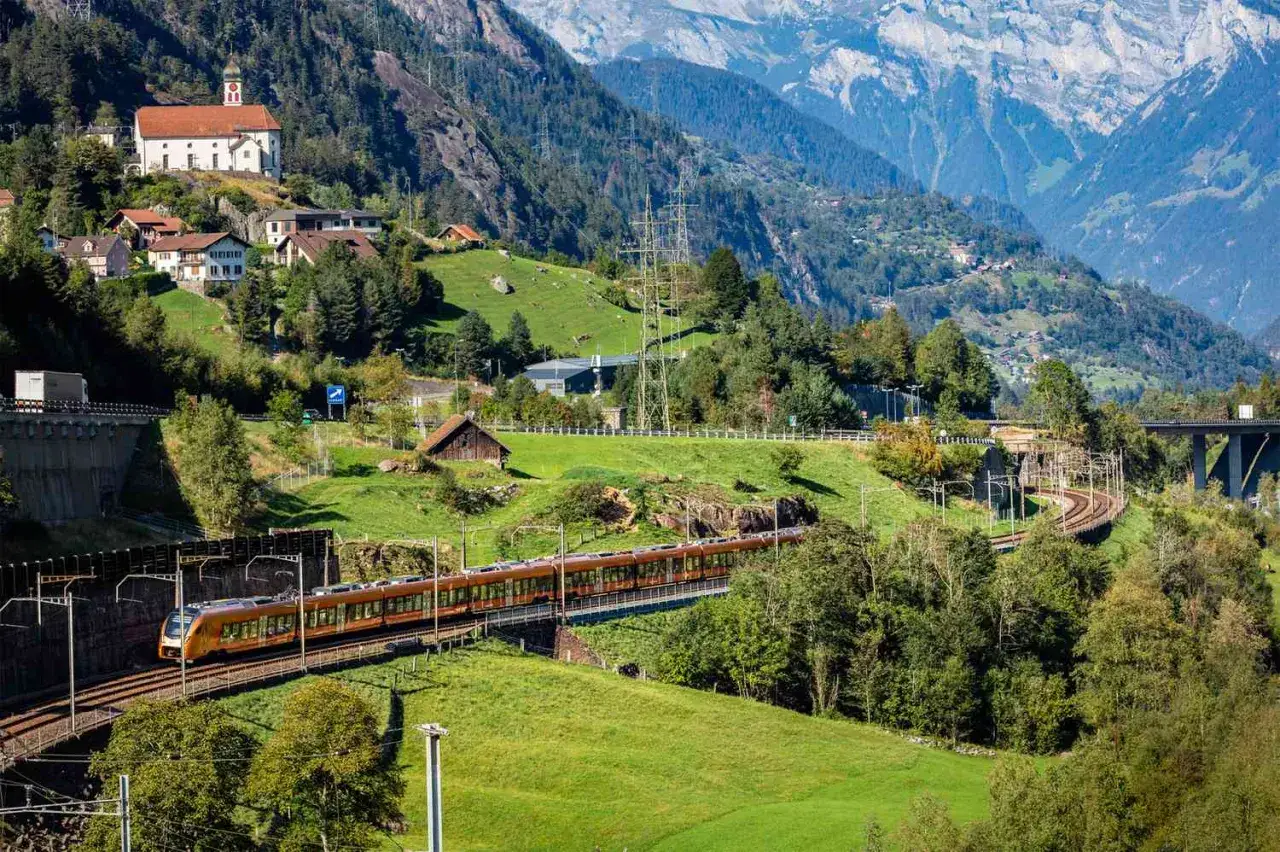 The Gotthard Panorama Express train winds through a lush green valley in the Swiss Alps, passing a charming village and a church.